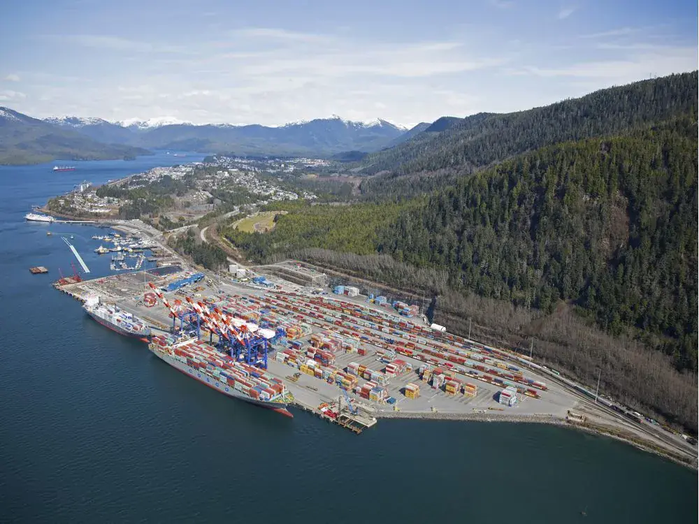 Aerial view of Fairview Container Terminal at the Port of Prince Rupert on Kaien Island, BC — a container ship at berth with gantry cranes, stacked containers, and the on-dock CN rail line following the shoreline below the Coast Mountains